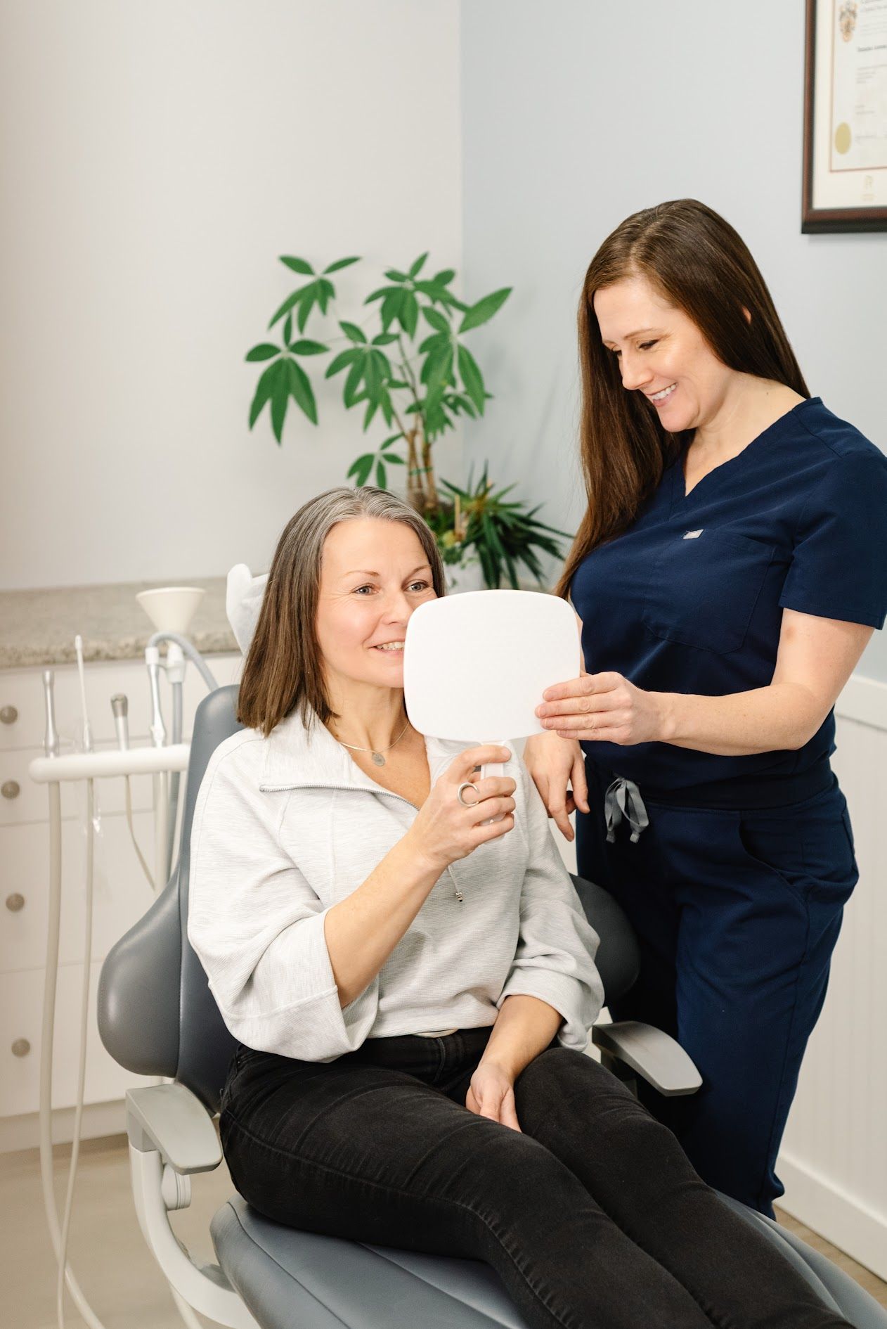 A healthcare provider in blue scrubs holds a clipboard, smiling while consulting with a patient in a light-colored top.