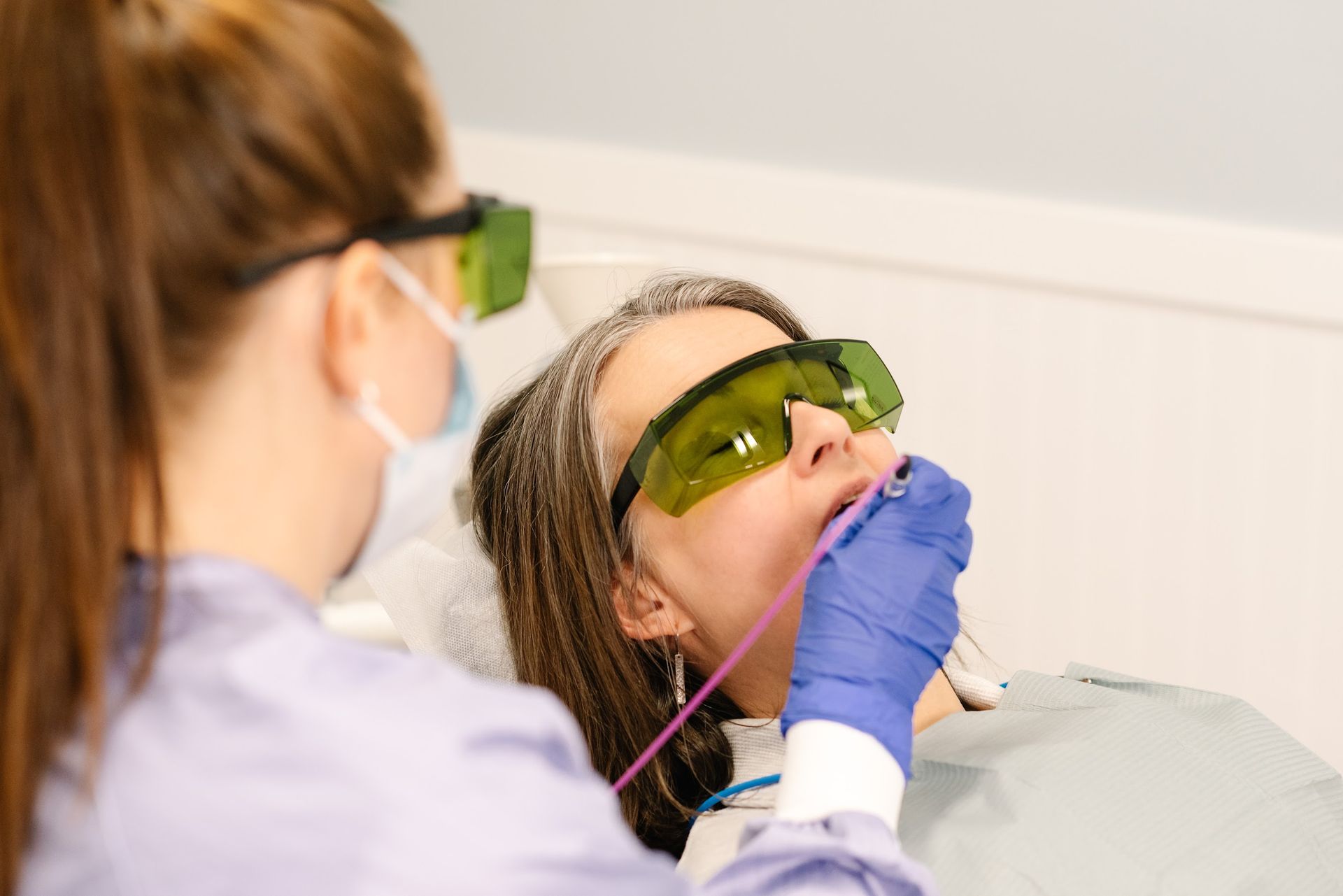 A medical professional in protective glasses performs a dental laser procedure on a patient wearing safety goggles.