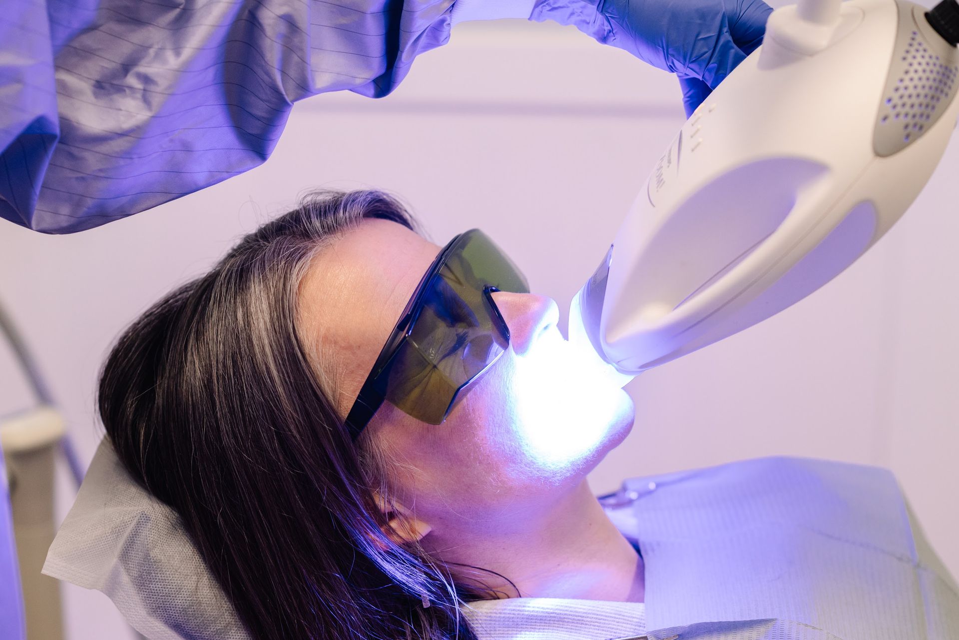 A patient wearing protective eyewear receives professional teeth whitening treatment with a blue light lamp in a clinic.