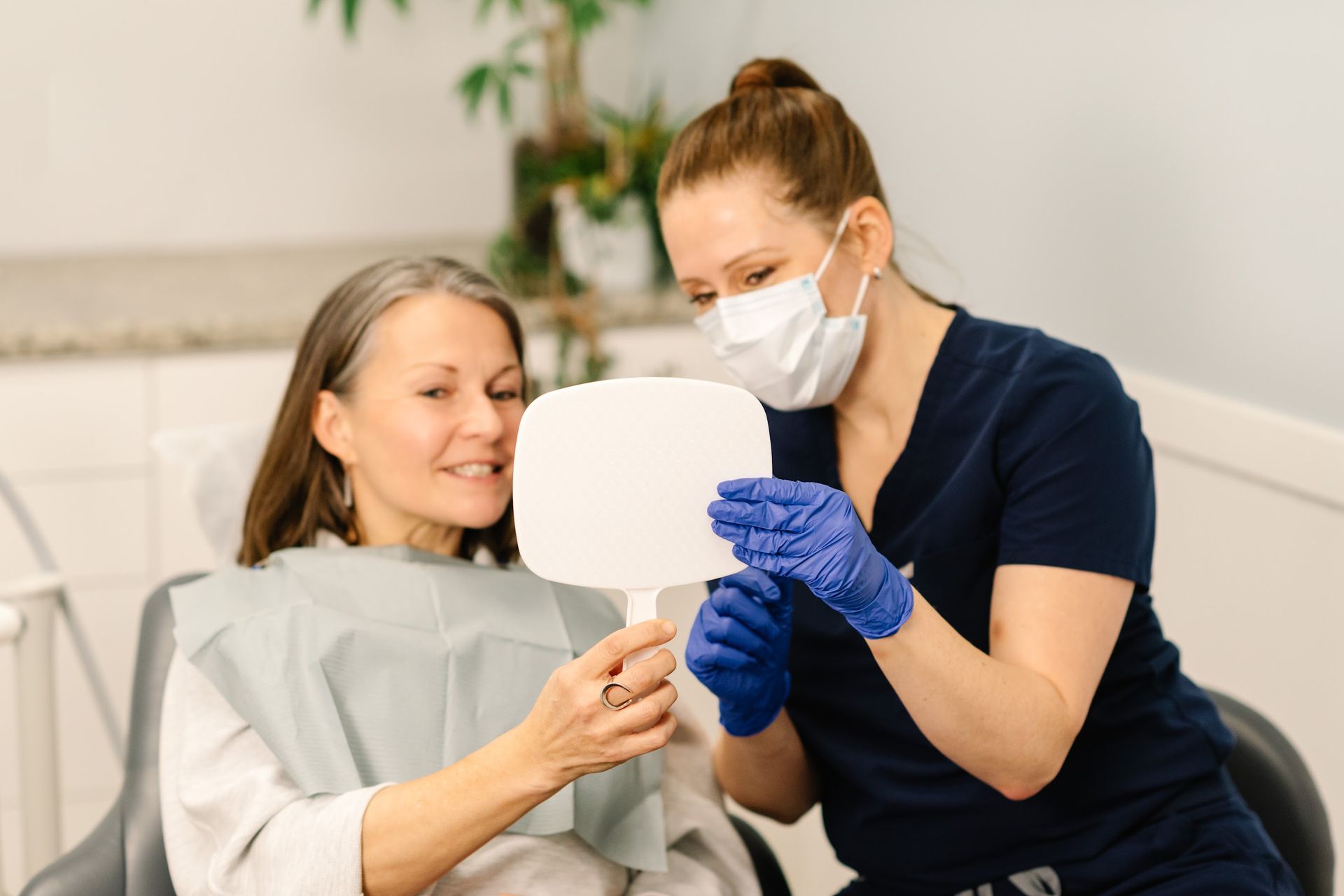 A dental professional wearing gloves and a mask shows a patient their results in a hand mirror inside a clinic.