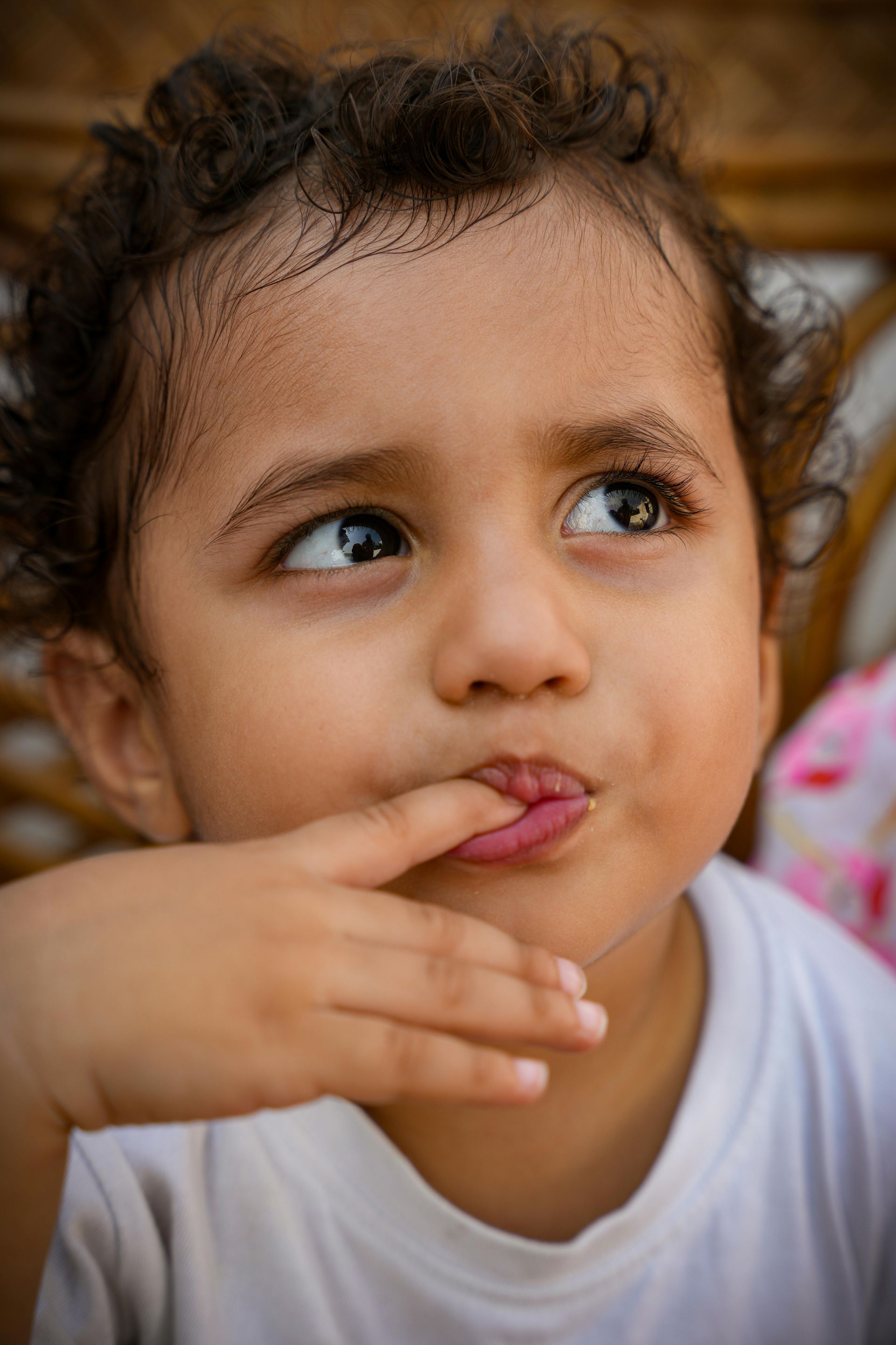 Young child with curly hair looking up thoughtfully, finger near lips, against a warm blurred background