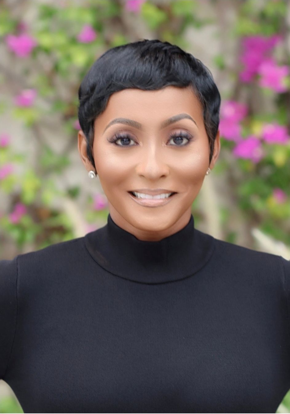 Black woman with arms crossed, smiling, wearing a grey blazer and peach top.