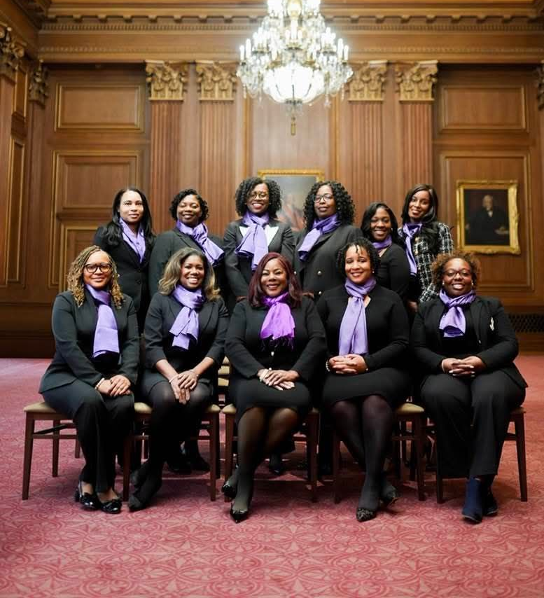 Group of Black women pose in front of a banner with a logo. Hallway setting.