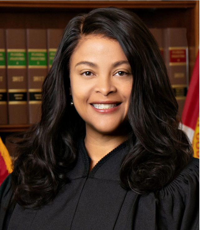 Judge smiling, wearing judicial robe, in front of law books and the Florida flag.
