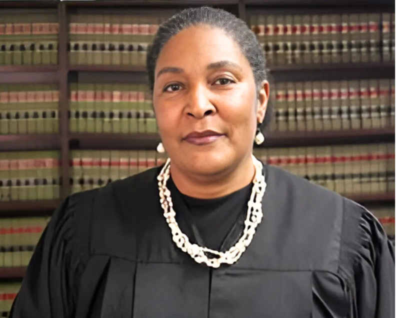 Black woman judge in black robe, pearl necklace, in a courtroom library setting.