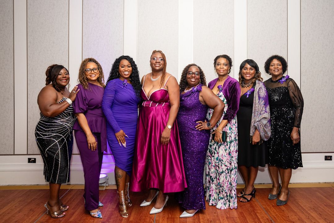 Group of people posing, mostly women in black outfits with purple scarves, a man in a suit, at an event.