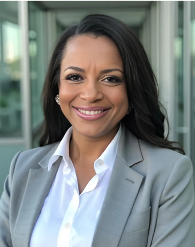 Woman with dark hair smiles in a grey blazer and white shirt, outdoors in front of glass.
