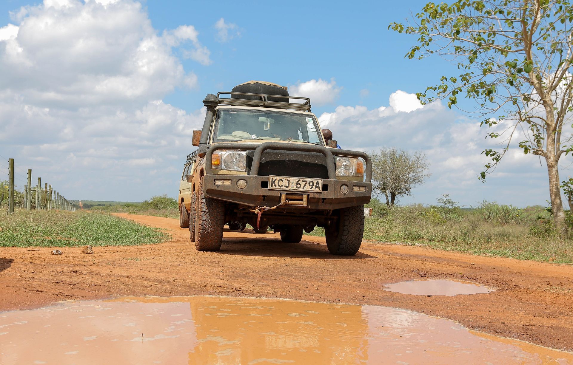 Mud-covered off-road truck driving on a dirt road with puddles under a partly cloudy sky