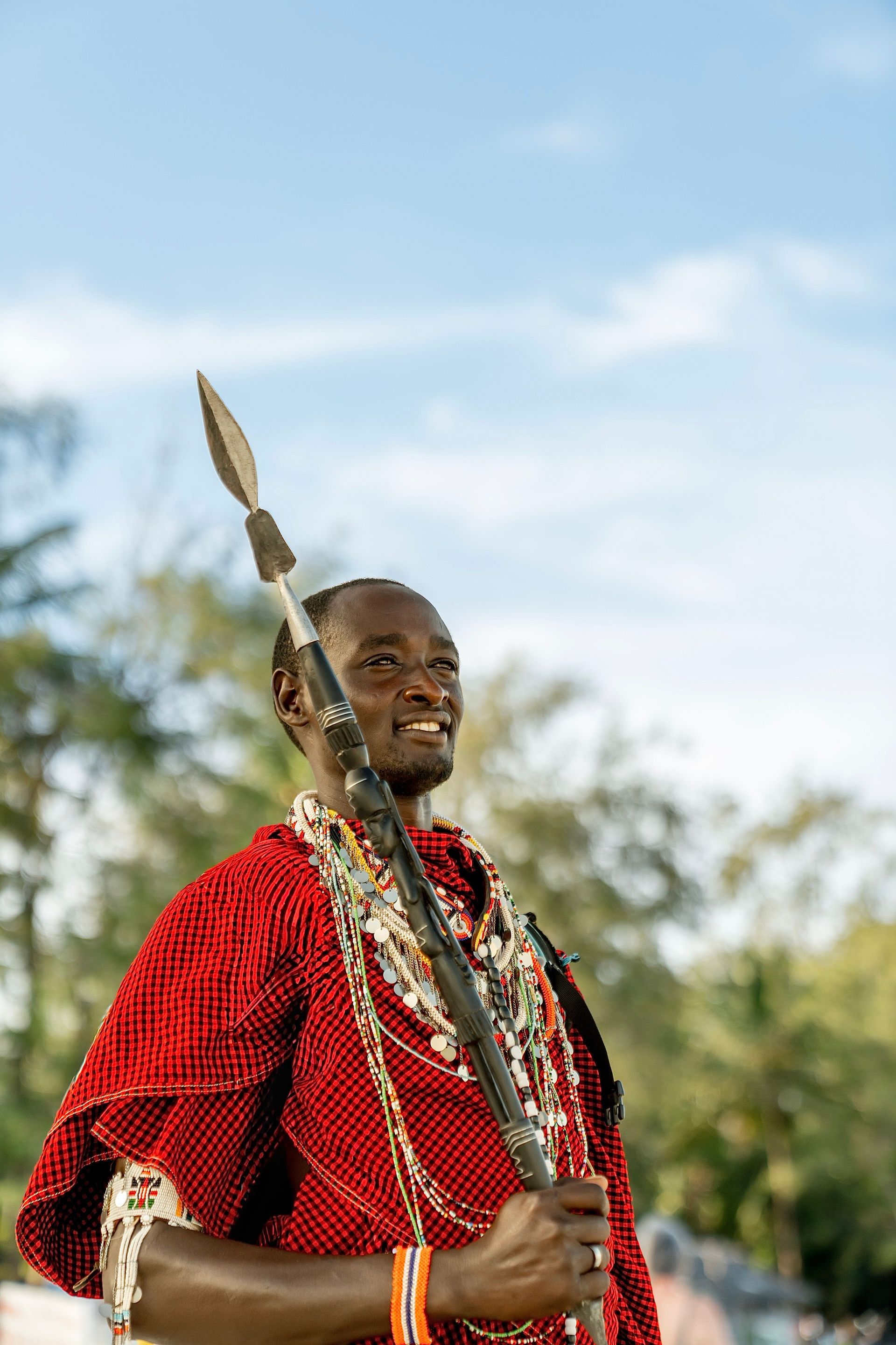 Man in a red beaded garment holding a spear outdoors under a blue sky