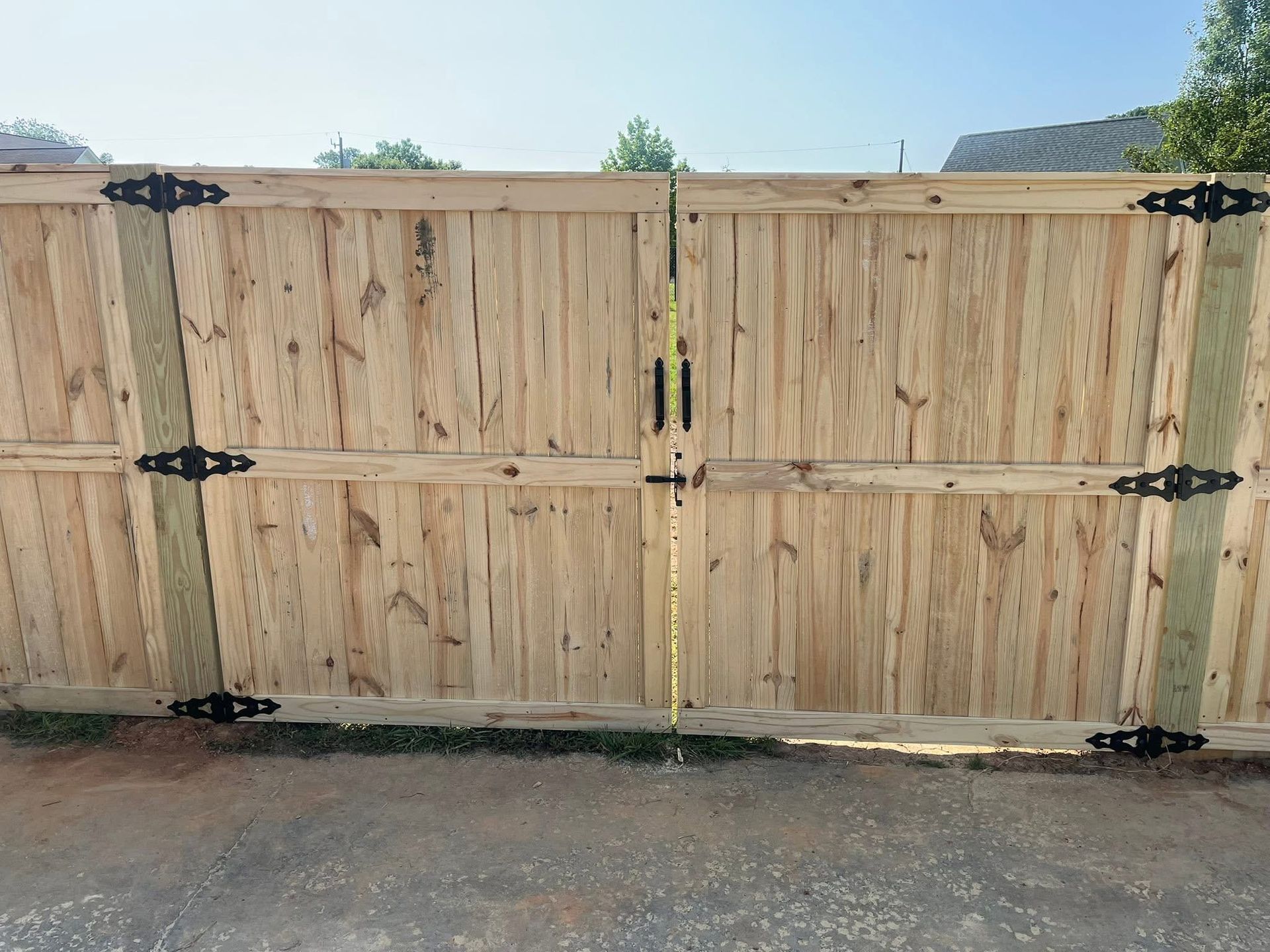 A double wooden gate with light-colored vertical planks and black ornamental hinges, set against a bright outdoor backdrop.