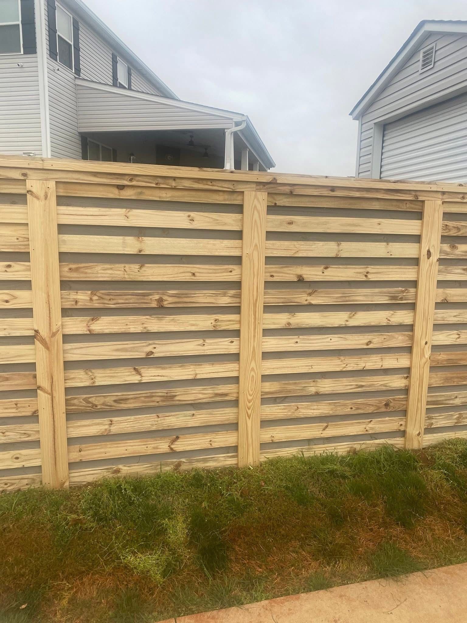A horizontal slat wood fence stands in front of two houses under a cloudy sky.