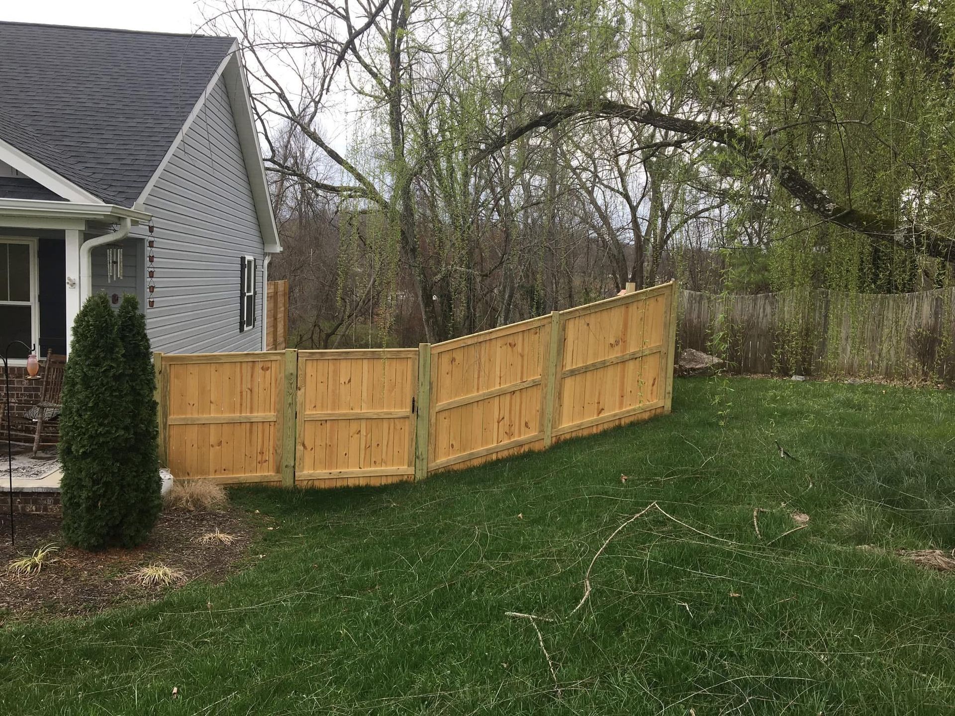 A light wood privacy fence runs from the corner of a gray siding house into a grassy backyard with trees.
