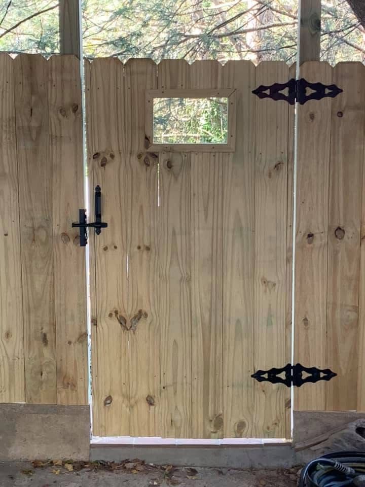 A wooden gate with black metal hinges and a latch, featuring a small rectangular window cutout, set in a fence.
