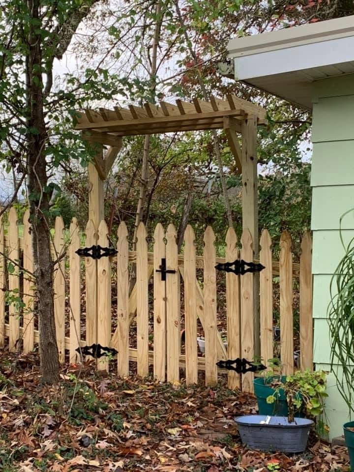 A wooden gate with a small pergola topper, featuring black hinges and a latch, set in a yard with trees and fallen leaves.