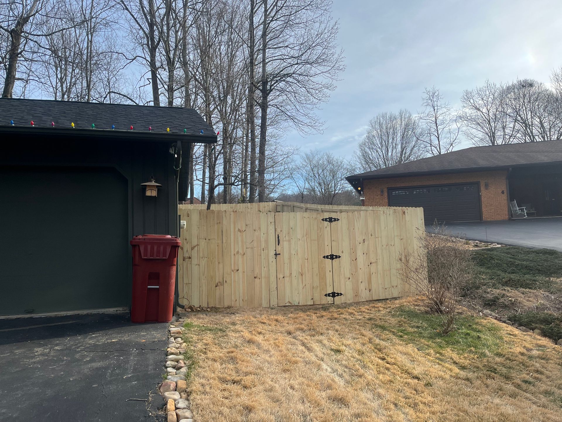 A wooden gate sits between a dark house and a grassy yard, next to a red trash bin on a driveway.