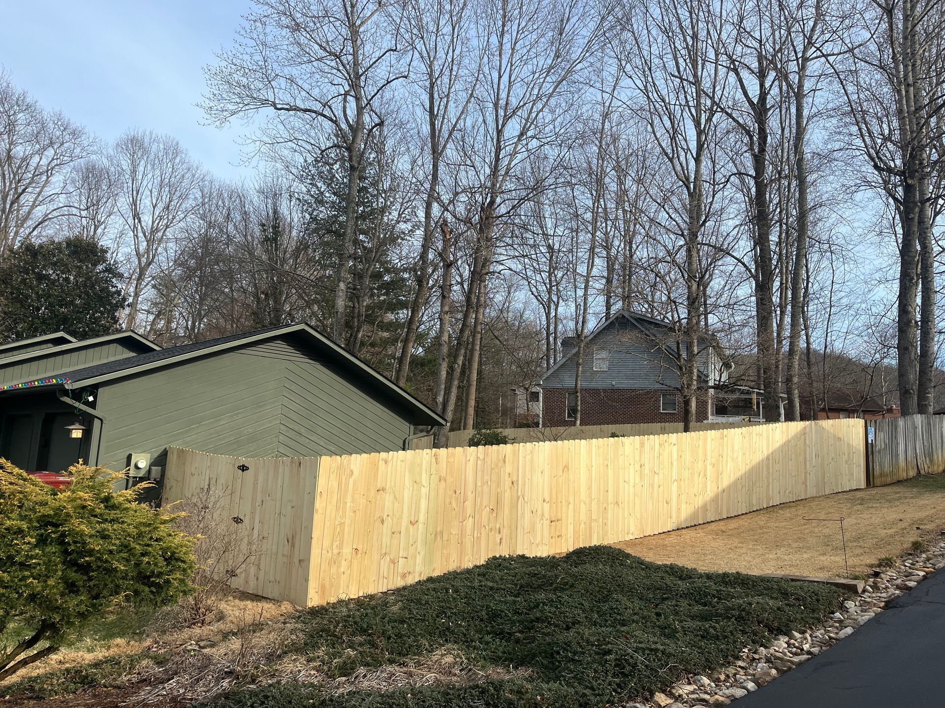 A long, newly installed light-wood privacy fence stands in a grassy yard in front of a house with olive-green siding.