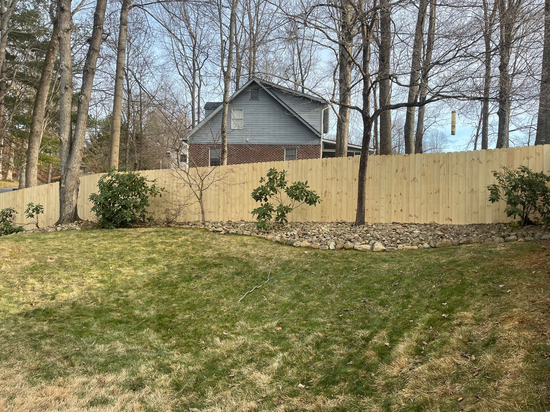 A light wood privacy fence stands in a grassy backyard, backed by tall trees and a partial view of a neighboring house.