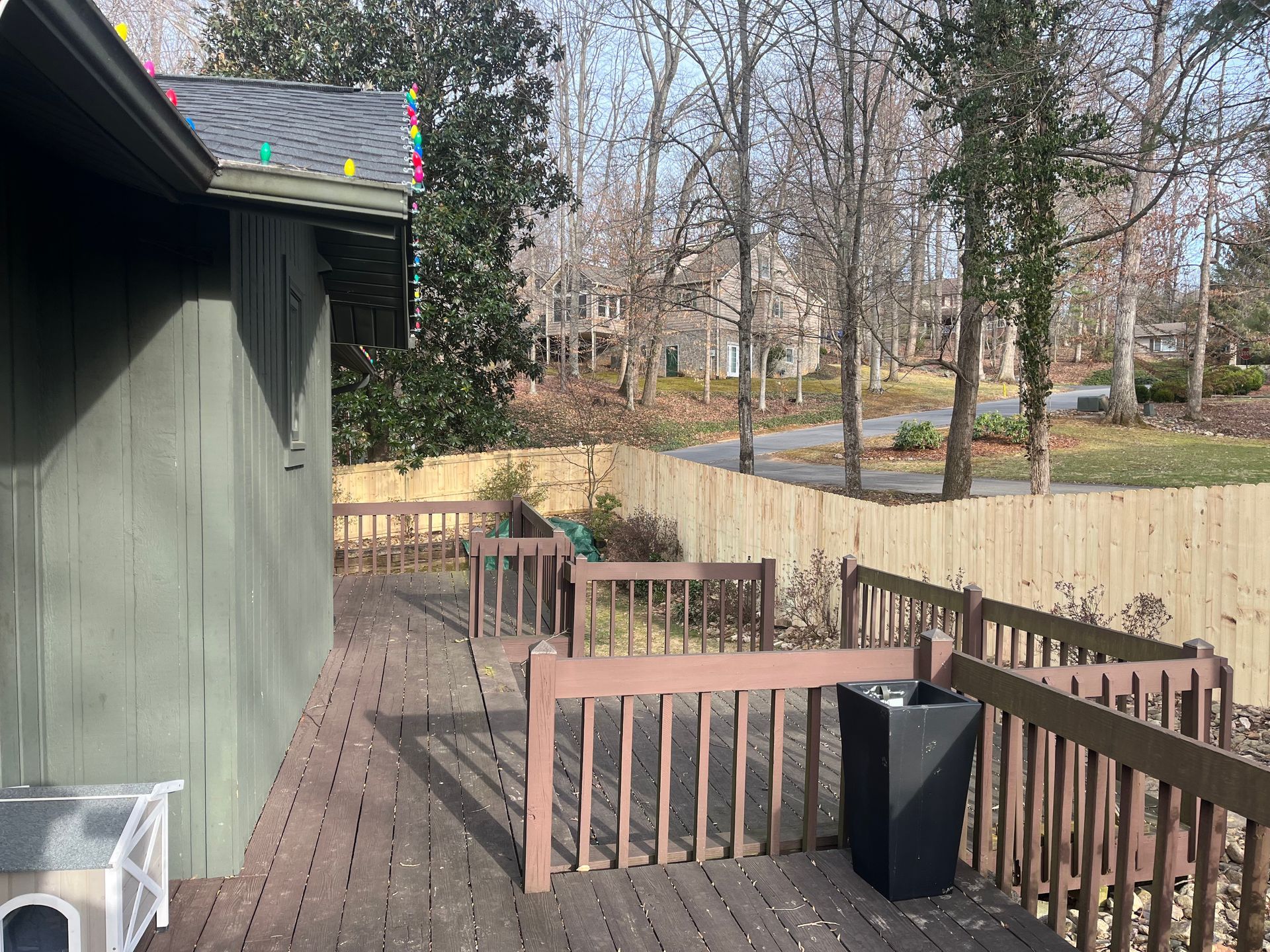 A wooden deck attached to a green house, with railings overlooking a fenced backyard area and trees in the distance.