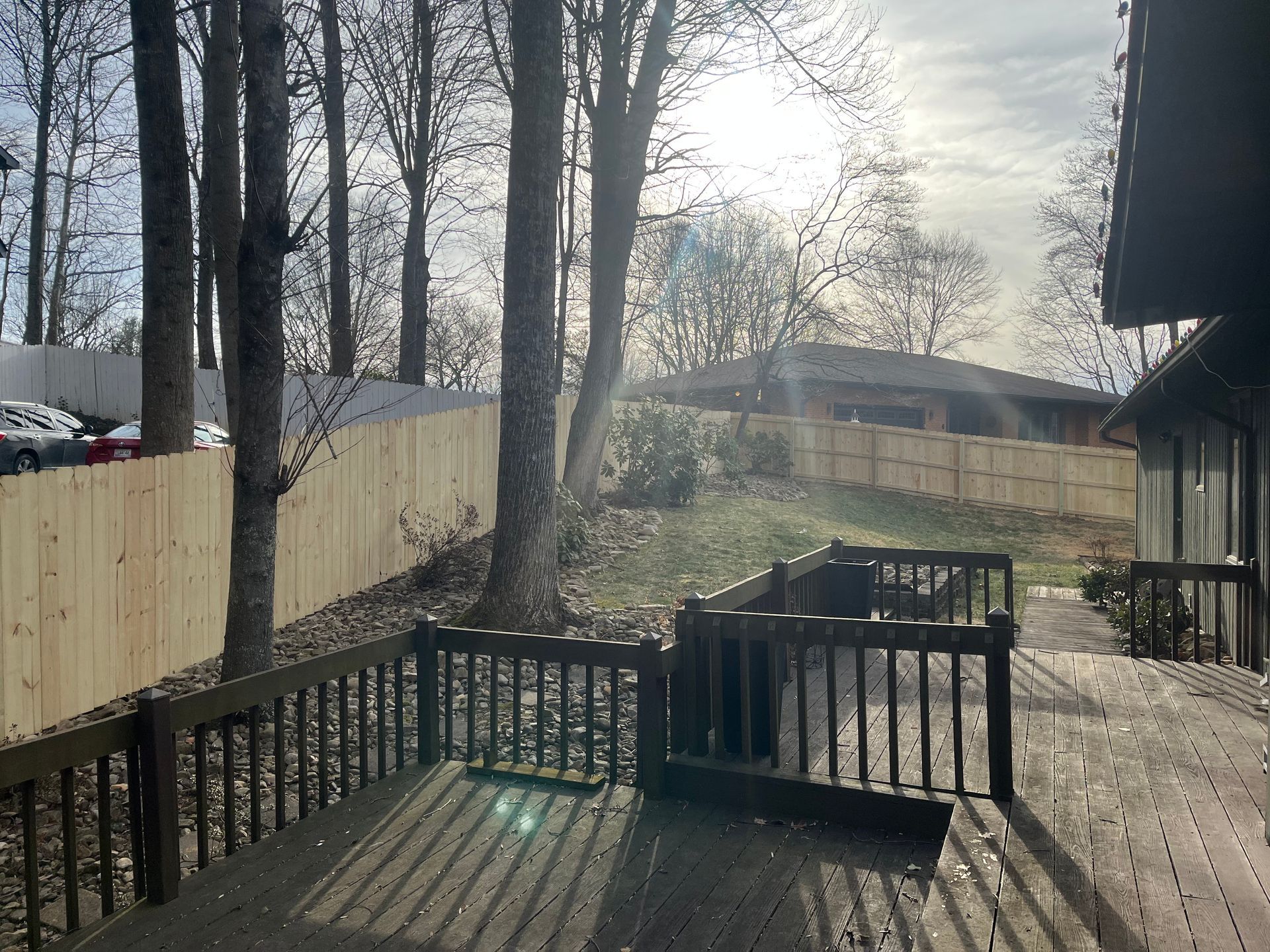 A wooden deck overlooks a grassy backyard enclosed by a new light-colored wood privacy fence under bright sunlight.