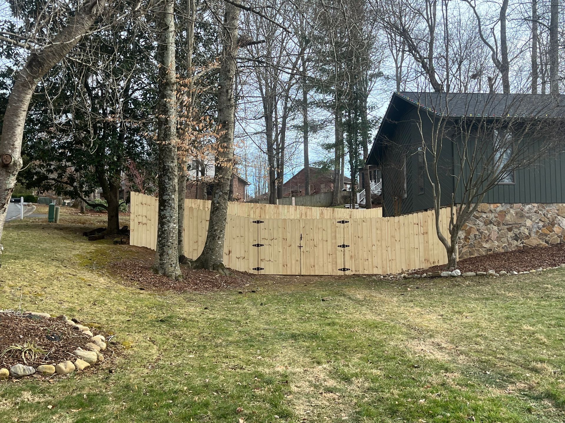 A new light-wood fence stands in a grassy backyard next to a dark house with a stone base and surrounding trees.