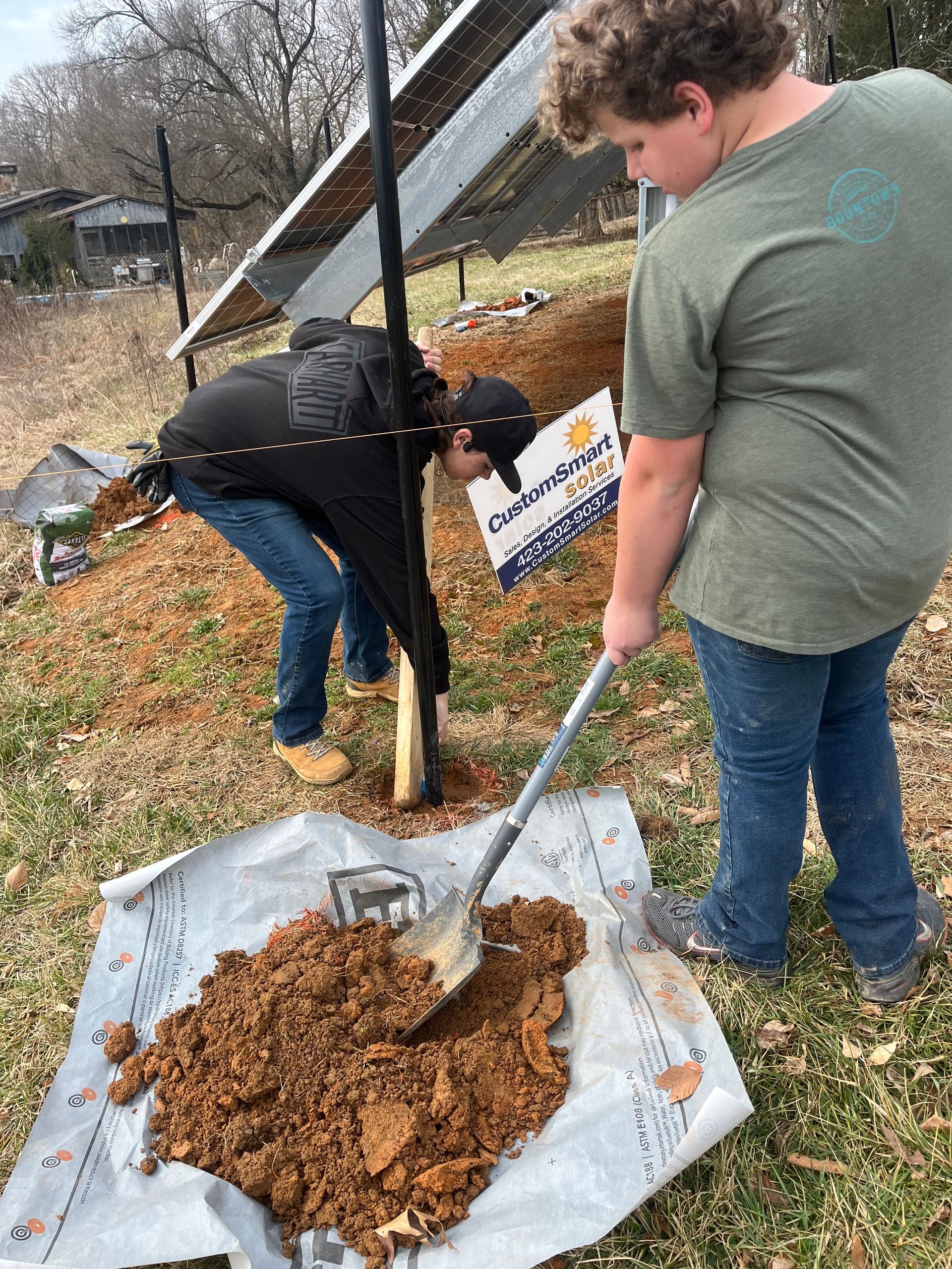 Two people use a shovel to move a pile of dirt onto a tarp outdoors near a solar panel structure.
