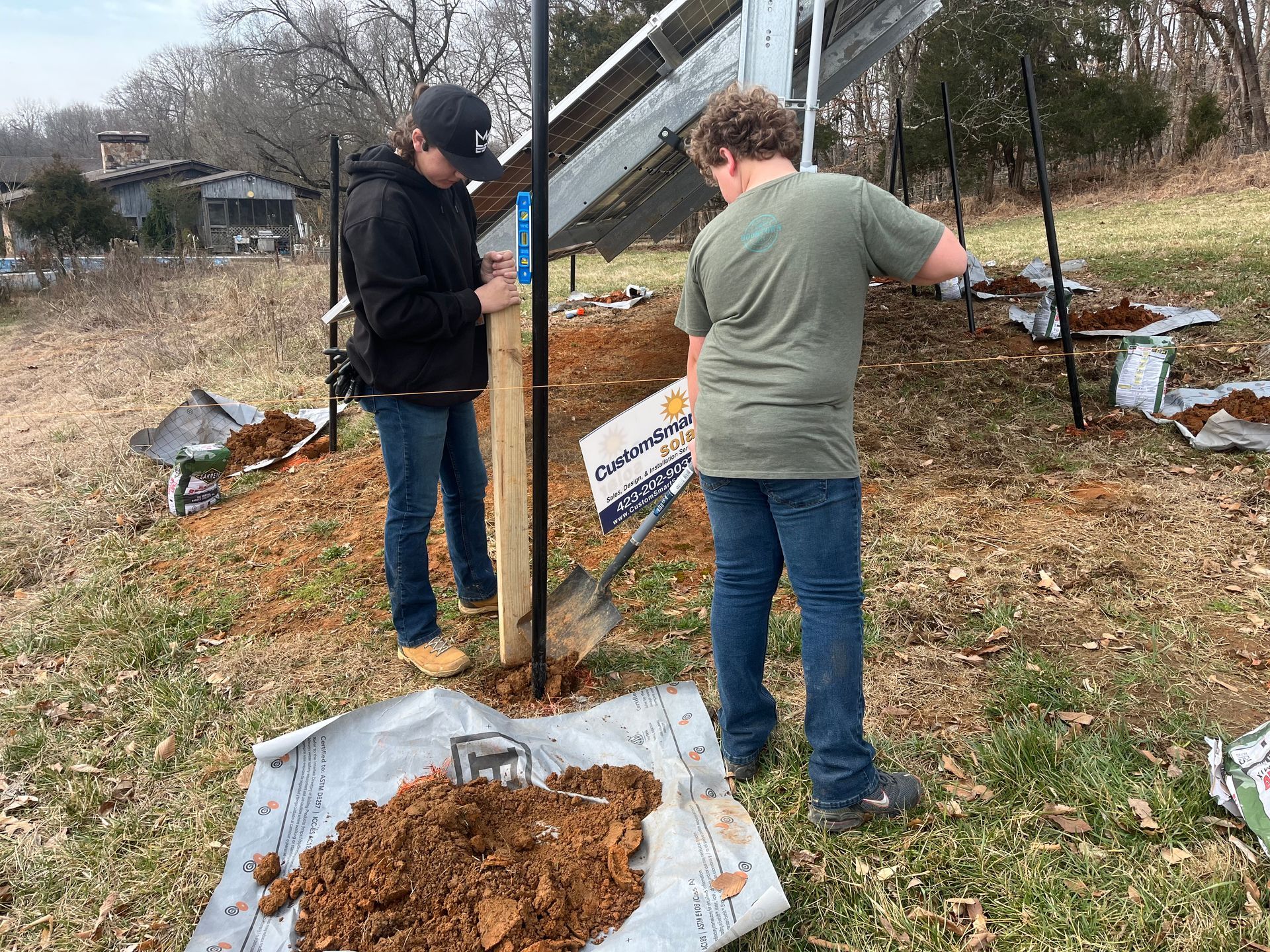 Two people in casual clothes dig holes for posts near a solar array in a grassy, outdoor area.