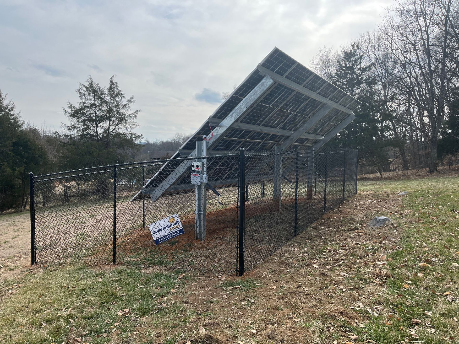 A tilted solar panel array enclosed by a black chain-link fence in a grassy, treed area on a cloudy day.