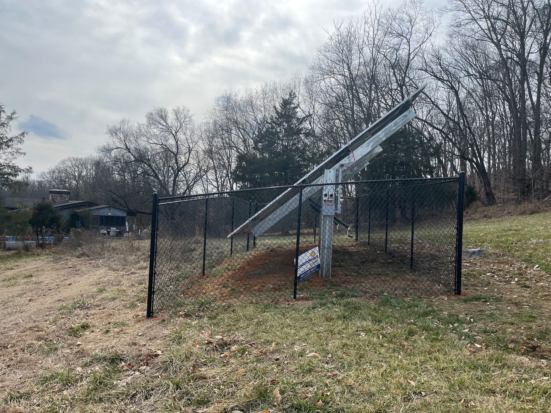 A solar panel array angled toward the sun, enclosed by a chain-link fence in a grassy, wooded outdoor area.