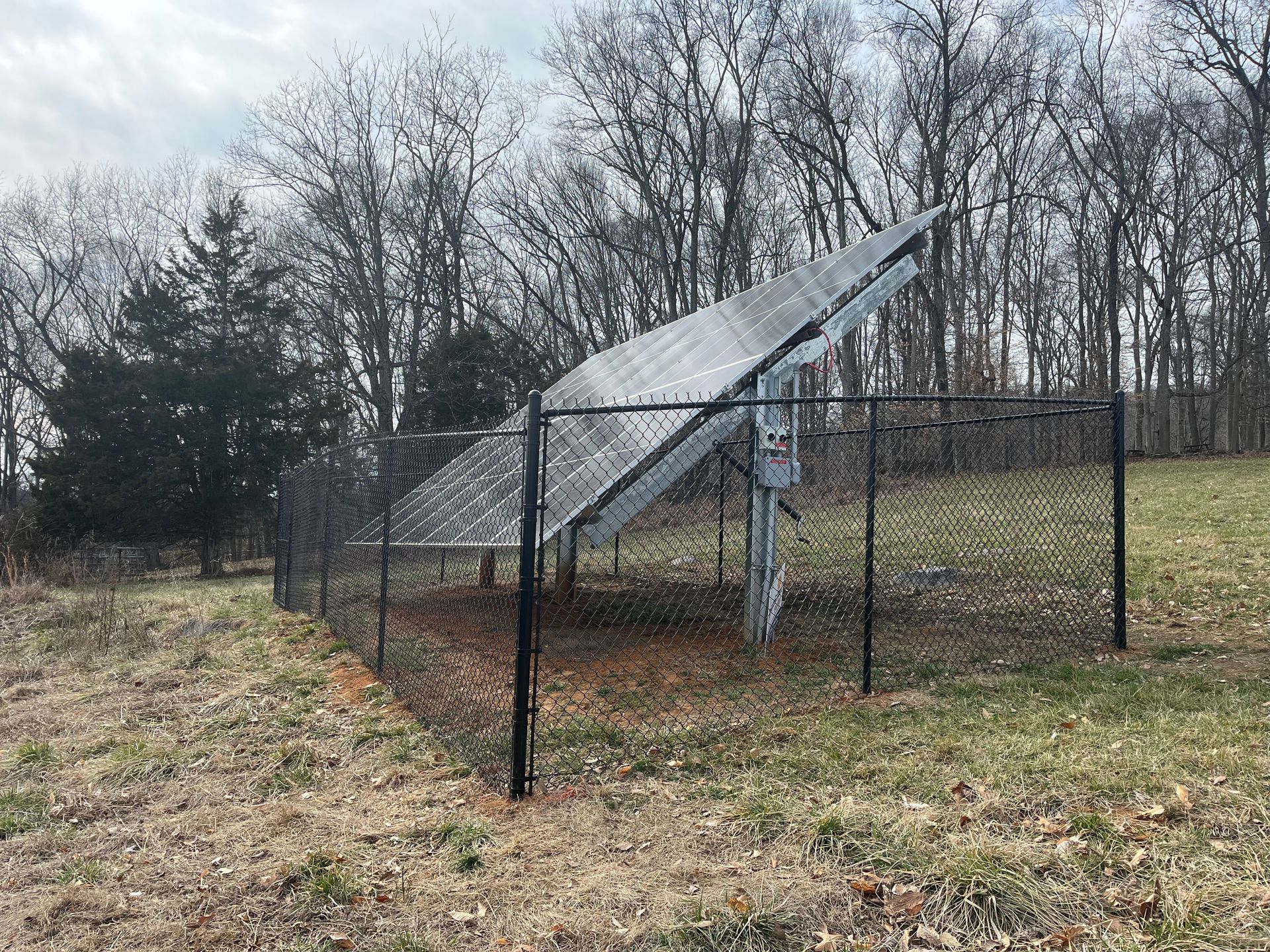 A small solar panel array mounted on a single post inside a chain-link fence, situated in a wooded, grassy field.