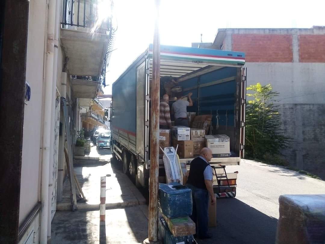 Un camion scarica pacchi in una strada stretta. Due persone dentro il camion, un uomo fuori. Giornata di sole.