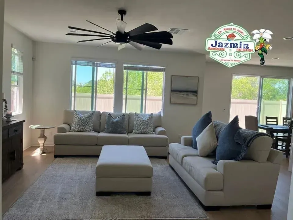 Bright living room with gray sofas, ottoman, large windows, ceiling fan, and light neutral decor
