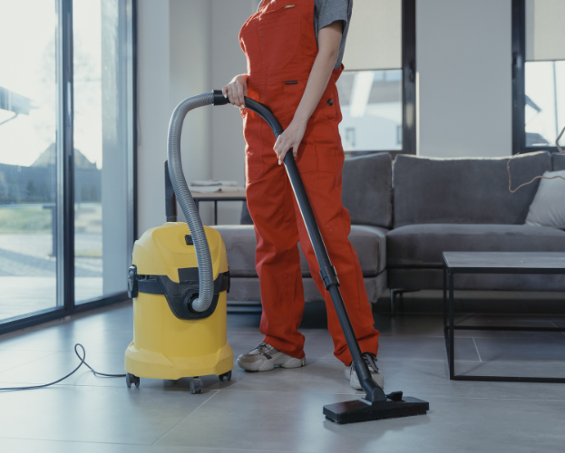 Person vacuuming a bright living room beside a yellow canister vacuum and gray sofa.