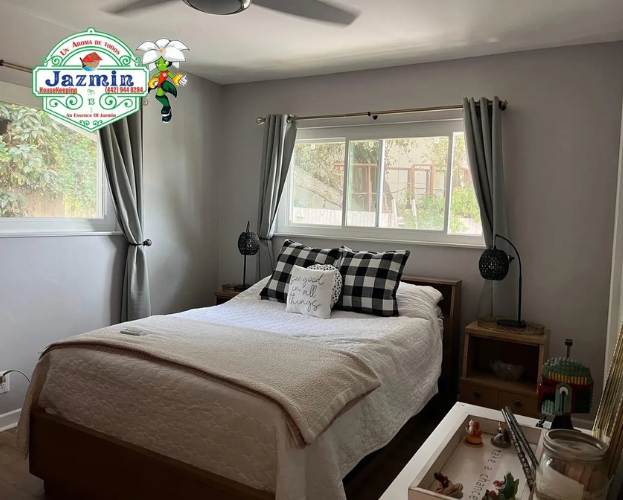 Bright bedroom with a neatly made bed, gray curtains, and a window letting in daylight.