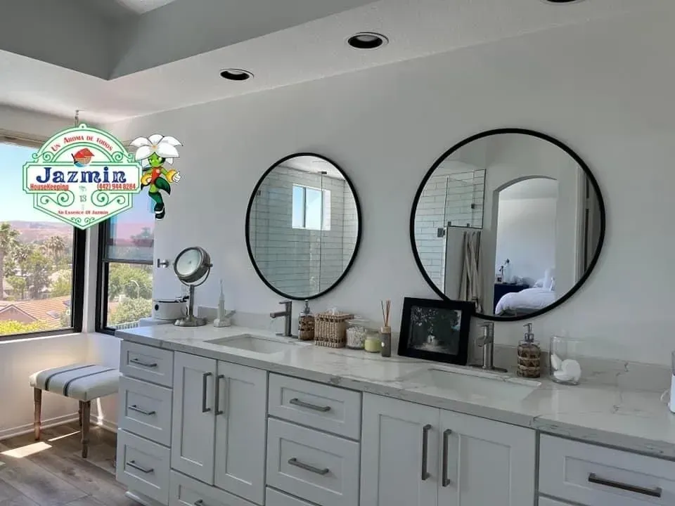 Bright bathroom vanity with two round mirrors, gray cabinets, and a window seat beside large windows.