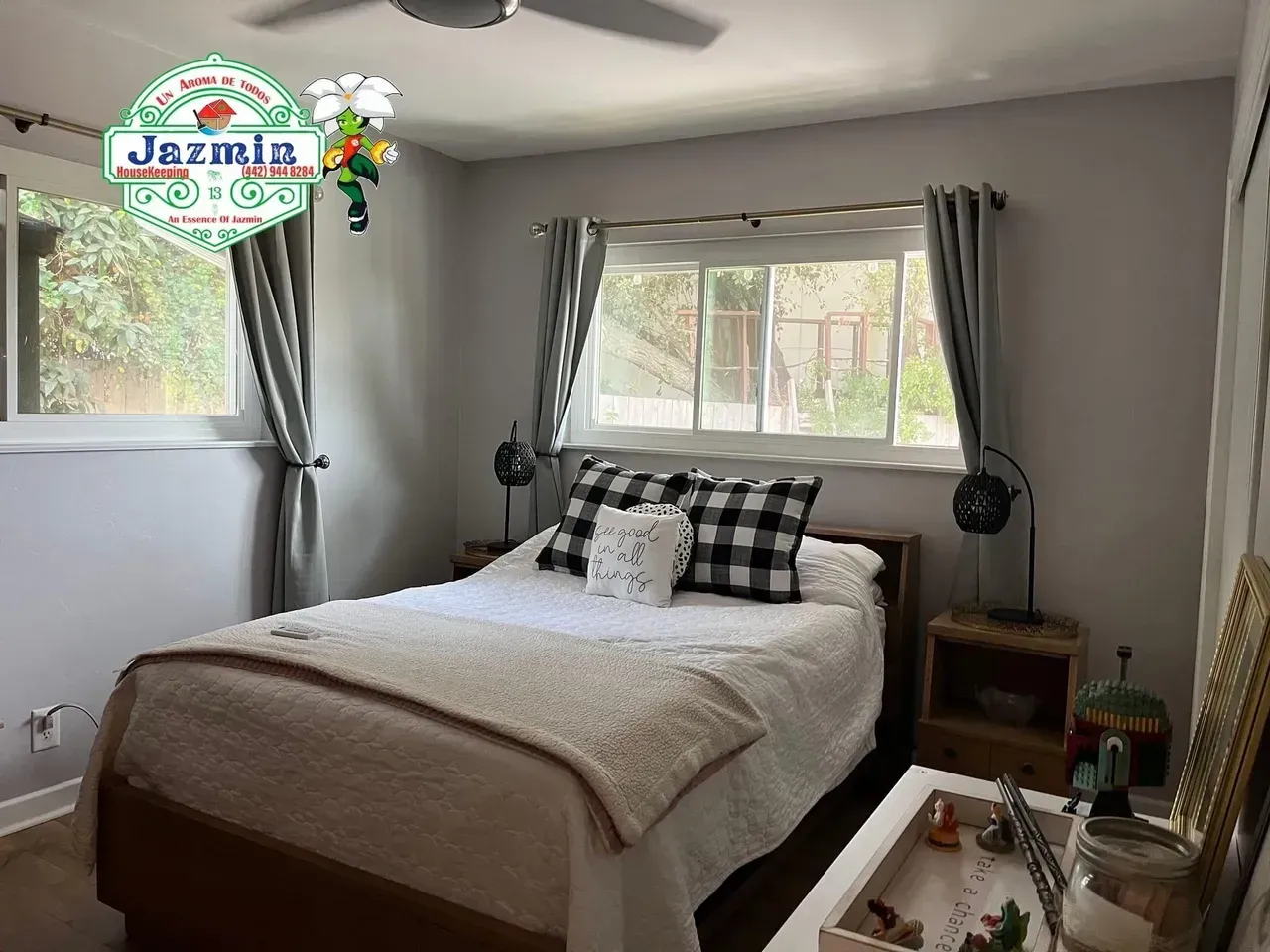 Bright bedroom with a made bed, plaid pillows, two windows, and a small bedside table.