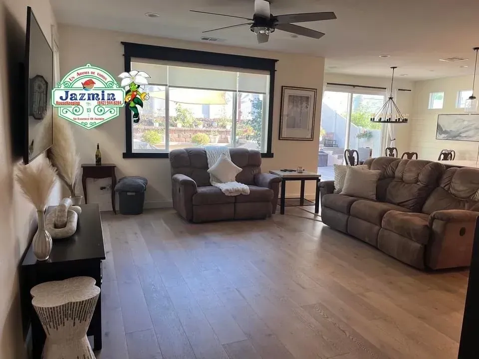 Bright living room with tan sofas, large window, ceiling fan, and wood-look flooring