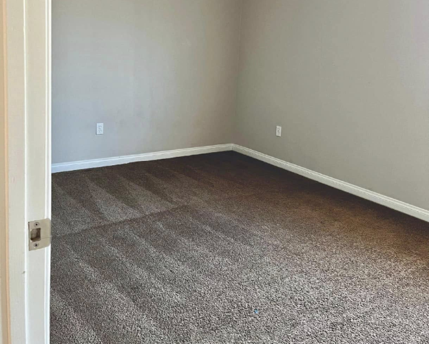 Empty beige room with brown carpet and white trim, viewed from the doorway.