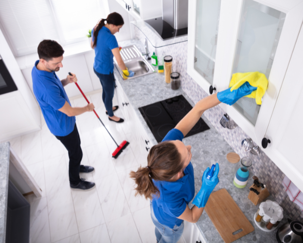 Three cleaners in blue uniforms cleaning a modern kitchen with mops, cloths, and spray bottles.