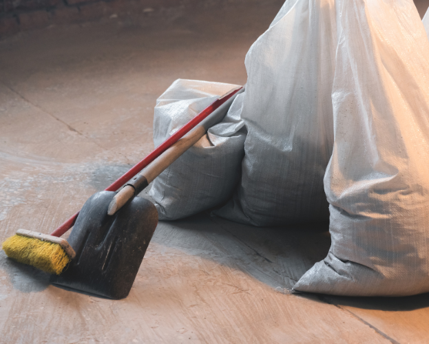 Broom and dustpan leaning against gray trash bags on a wooden floor