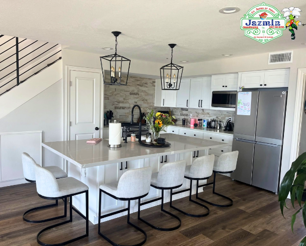 Modern white kitchen with island, white barstools, pendant lights, stainless fridge, and dark wood floors