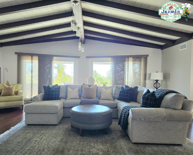 Bright living room with a gray sectional, round ottoman, exposed beams, and large windows.