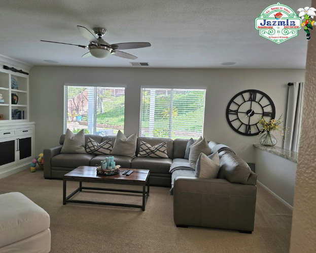 Bright living room with gray sectional, coffee table, wall clock, window blinds, and ceiling fan