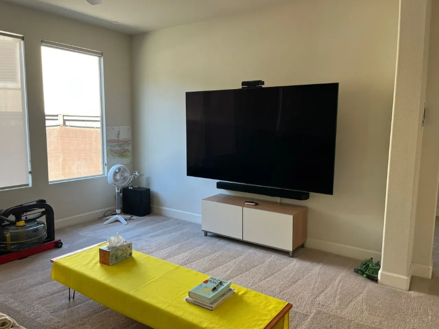 Bright living room with a wall-mounted TV, yellow coffee table, and large window.