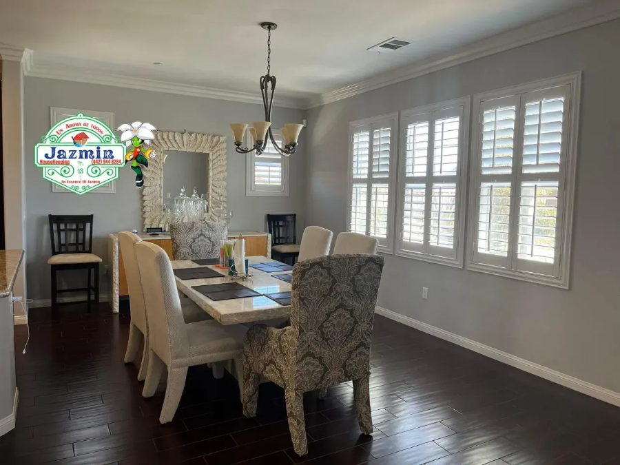 Dining room with a wooden table, upholstered chairs, chandelier, and large white plantation-shutter windows.
