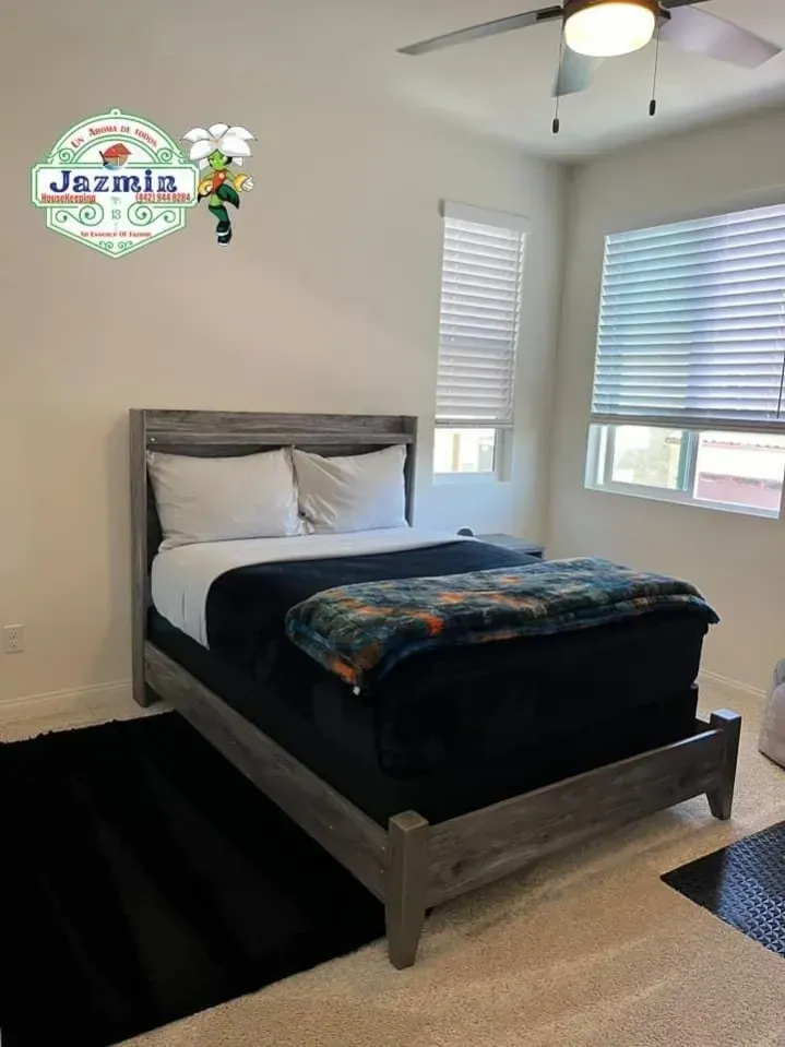 Small bedroom with a gray bed, black bedding, white walls, and two windows with blinds.