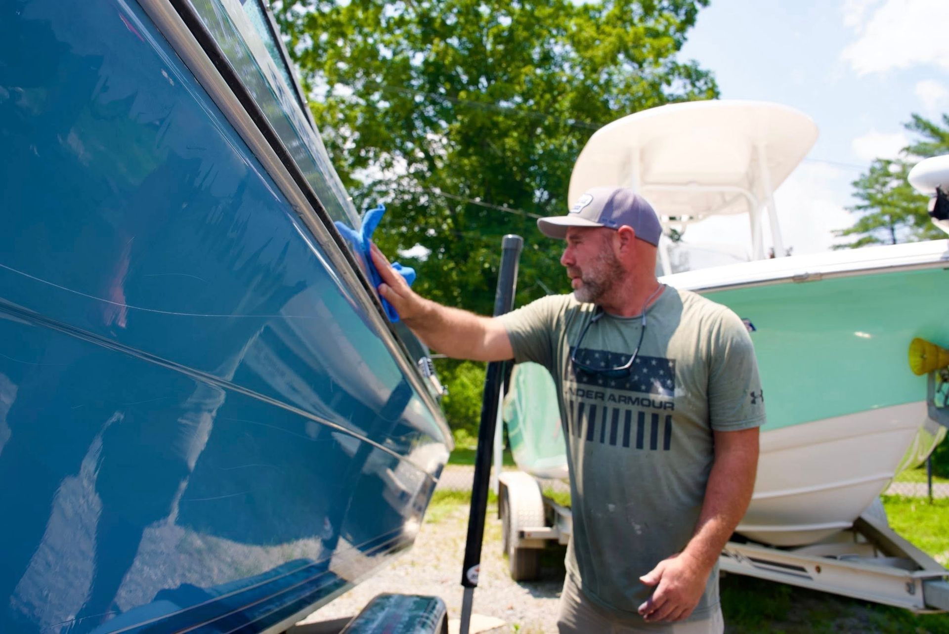 Man spray-painting a blue boat outdoors beside a white and green boat trailer.