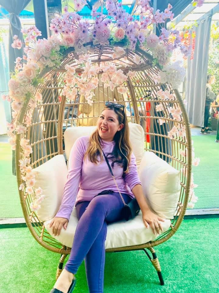 Woman in purple clothes sitting in a hanging chair decorated with flowers, sticking out tongue.
