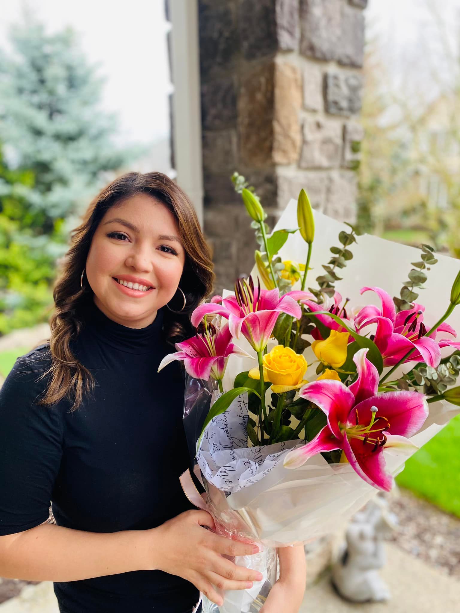 Woman smiling, holding bouquet of pink lilies and yellow roses; front of a house.