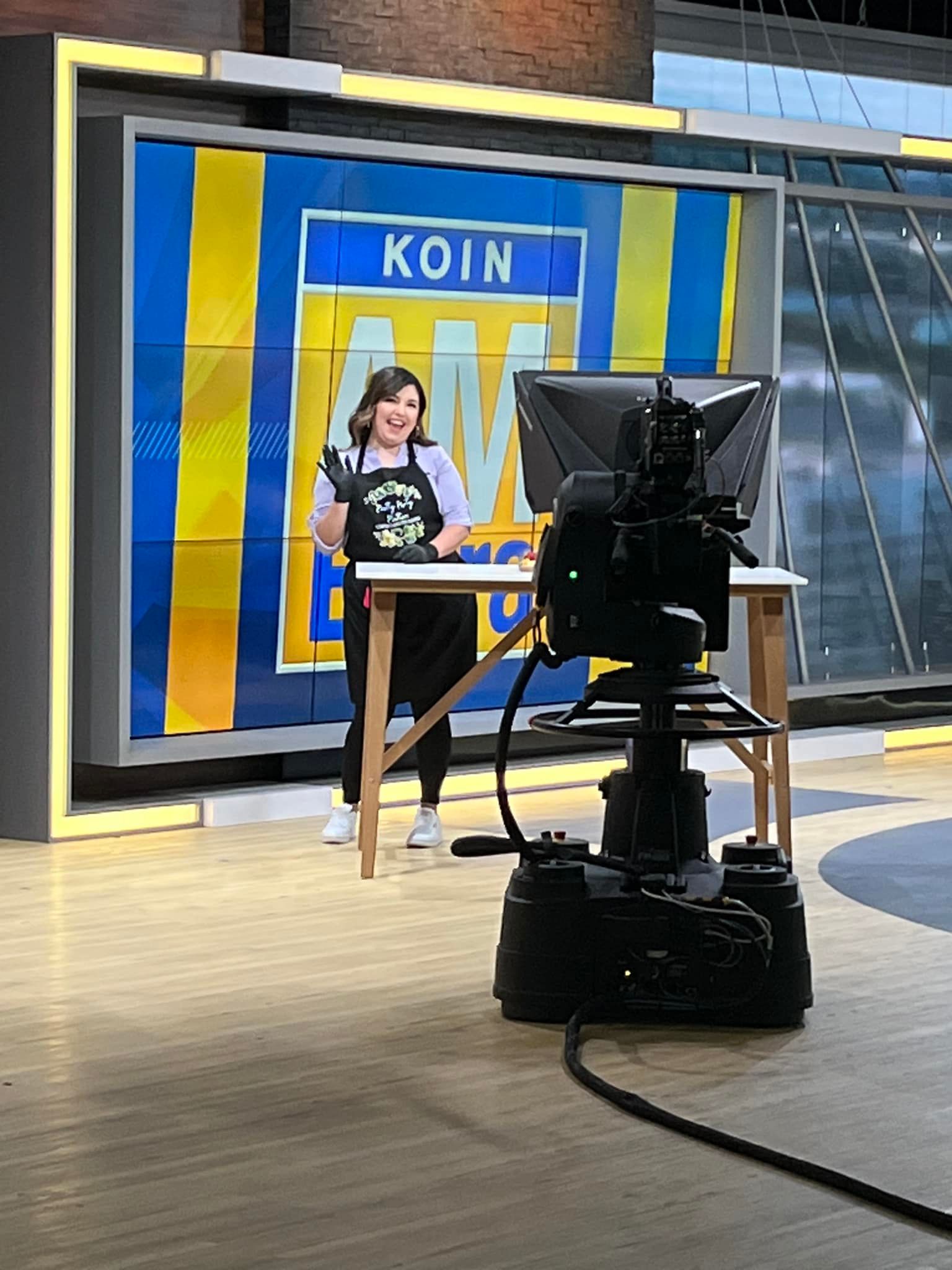 Woman on a TV set with a table, holding a microphone, smiling. KOIN logo on a blue and yellow backdrop.