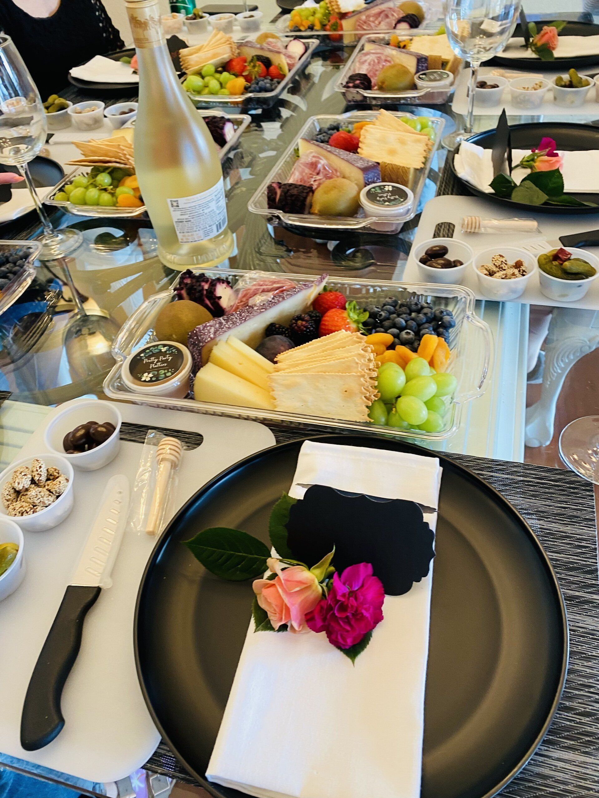 A table set with charcuterie boards, wine, and place settings. Floral napkin rings and various foods are featured.